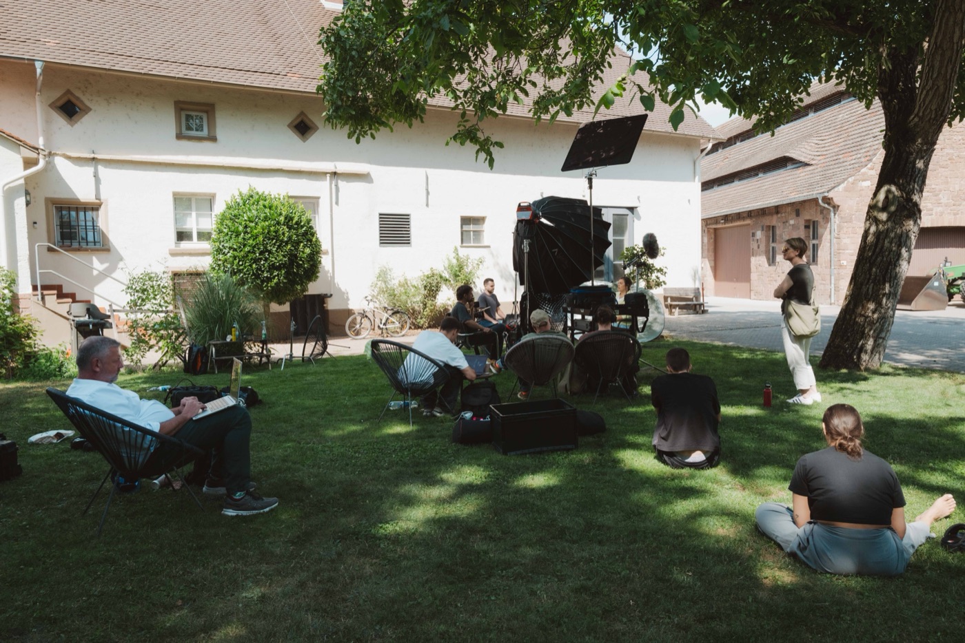 Dokumentarischer Außendreh: Crew und Monitore im Schatten eines Baums vor einem Bauernhaus.