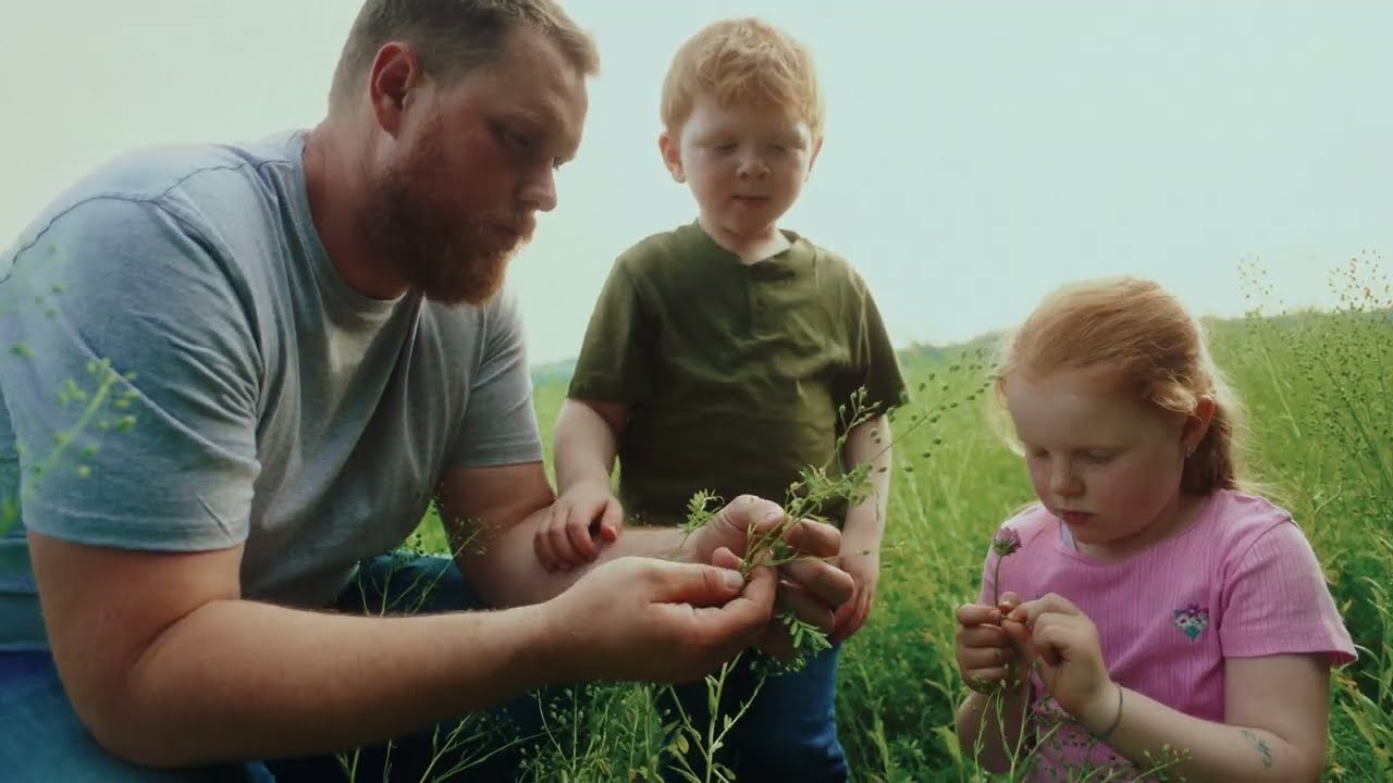 Standbild: Bio-Bauer hockt mit zwei Kindern im Feld und zeigt ihnen eine Pflanze.